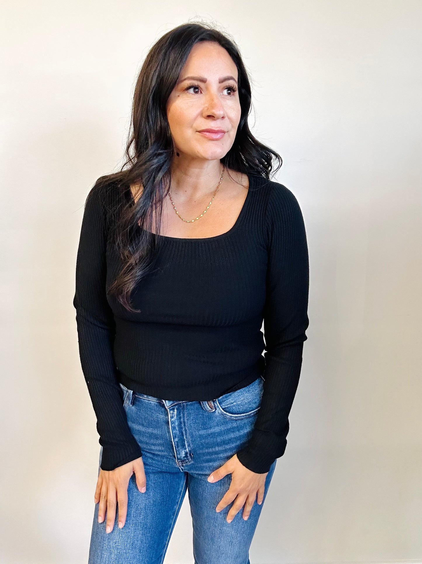 Woman wearing a black long-sleeve top and blue jeans against a plain background