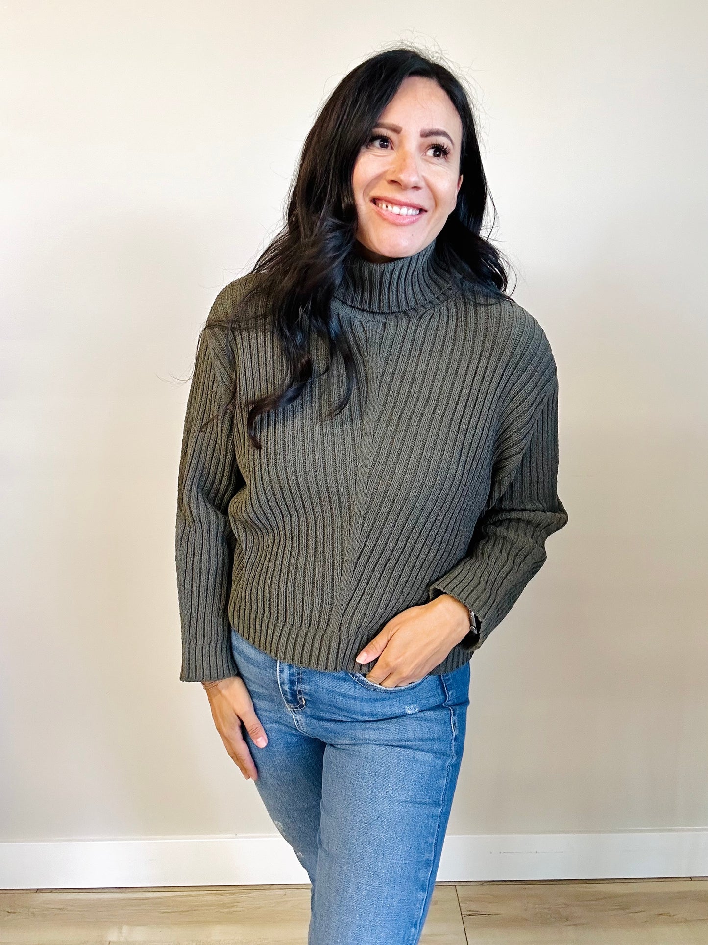 Woman wearing a dark green ribbed turtleneck sweater and blue jeans against a plain background