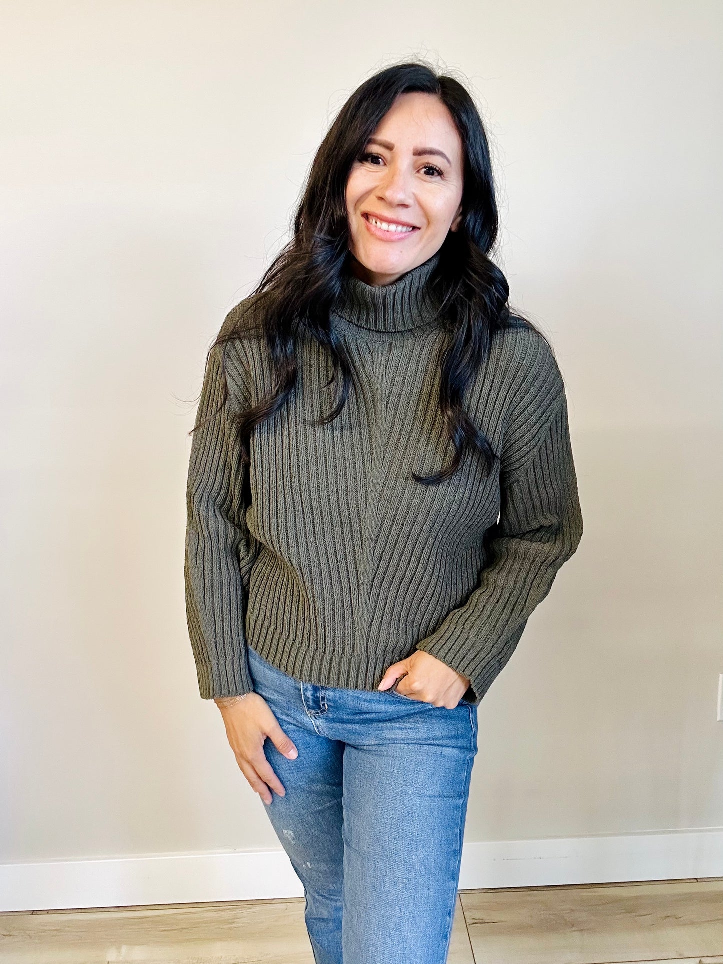 Woman wearing a green turtleneck sweater and blue jeans against a plain background